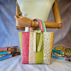 May include: A quilted fabric tote bag featuring vertical panels of various patterns including pink script, multicoloured dots, green floral, and yellow gingham. The bag has striped handles in red, green, yellow, and white, and a central fabric tie.