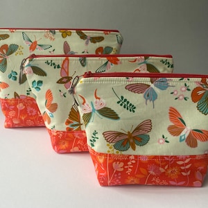 May include: Three zippered pouches with a white background and a colourful butterfly pattern. The pouches are lined with a red fabric with a floral pattern. The pouches are different sizes.