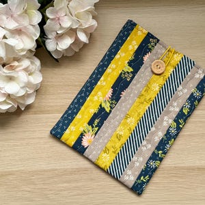 May include: A quilted book sleeve with a button fastening. The sleeve has vertical stripes in navy, yellow, grey, and white floral and striped patterns. A light-coloured hydrangea flower arrangement is in the background.