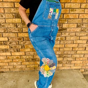 May include: Light blue denim overalls with decorative patches on the bib and knee. The bib pocket features colorful fabric patches, while the knee has a flower-shaped patch with a yellow center. The overalls have a classic design with adjustable straps.