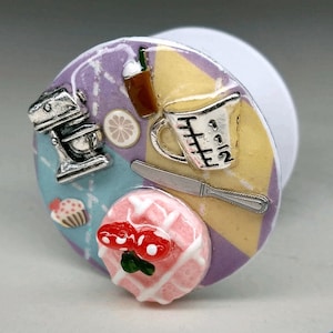 May include: A colorful phone grip with a pink and white waffle, a silver measuring cup, a silver mixer, a brown coffee cup, a lemon slice, and a silver knife.