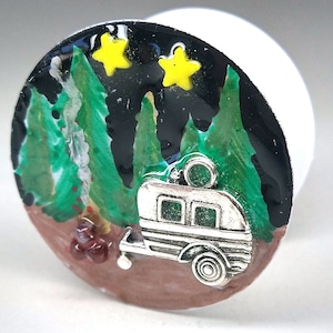 May include: A round, silver charm with a painted image of a camper van in a forest setting. The camper van is silver with black windows and a black roof. The forest is painted in shades of green and brown, with two yellow stars in the sky.
