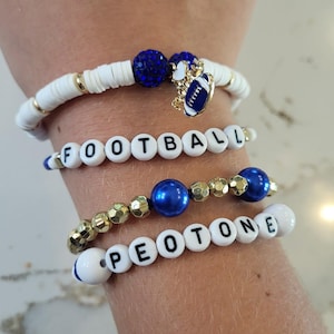 May include: Four beaded bracelets in blue, white, and gold. One bracelet spells "FOOTBALL" with letter beads. Another features a football charm. The bracelets are on a wrist.
