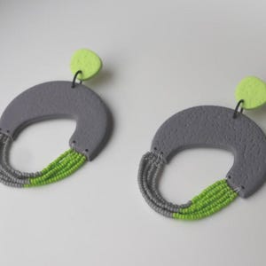 Può includere: Un paio di orecchini grigi, strutturati, a forma di semicerchio, con un gruppo di perline verdi nella parte inferiore e un piccolo accento verde lime nella parte superiore. Gli orecchini sono su sfondo bianco.