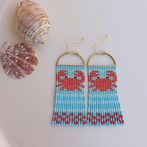 Puede incluir: Pendientes de cuentas con diseño de cangrejo. Los pendientes tienen un fondo a rayas azules y blancas con un cangrejo rojo. Acabados con ganchos dorados y flecos de cuentas. Un accesorio de verano.