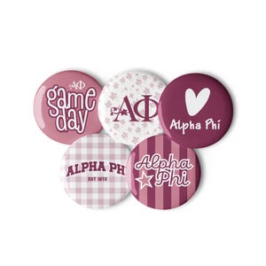 May include: A set of five circular pin-back buttons featuring maroon and white designs. The buttons display the text Alpha Phi, game day, and EST 1872, along with floral patterns, a heart, a star, plaid, and striped backgrounds in various shades.