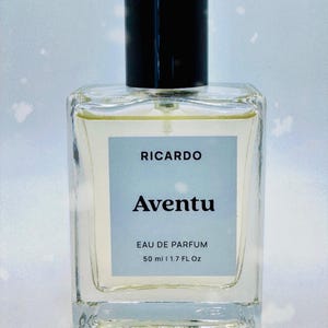 以下が含まれることがあります： 黒いキャップが付いた透明なガラスの香水瓶。「RICARDO Aventu EAU DE PARFUM 50 ml | 1.7 FL Oz」と書かれたラベル。中の液体は薄い黄色です。背景は水色です。
