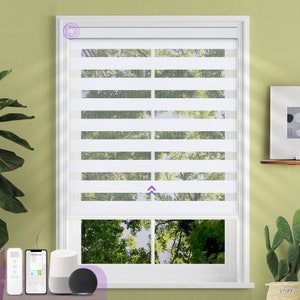 May include: White window blinds with a smart home device and a remote control. The blinds are raised to show a view of trees outside the window.