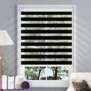 May include: Black motorised window blinds with a white frame. The blinds are partially raised, revealing a view of trees outside the window. The blinds are controlled by a remote control and a smart home device.