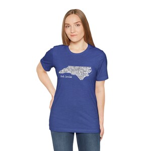Pu&ograve; includere: Una t-shirt blu reale con un contorno bianco dello stato della Carolina del Nord pieno di testo. Il testo include citt&agrave;, monumenti e altre cose associate allo stato. Il testo recita "Carolina del Nord".