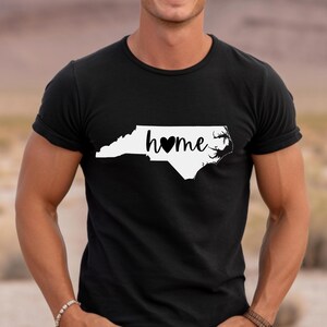 May include: Black t-shirt with a white outline of North Carolina and the word "home" with a black heart. The shirt is made of a soft material and has short sleeves.
