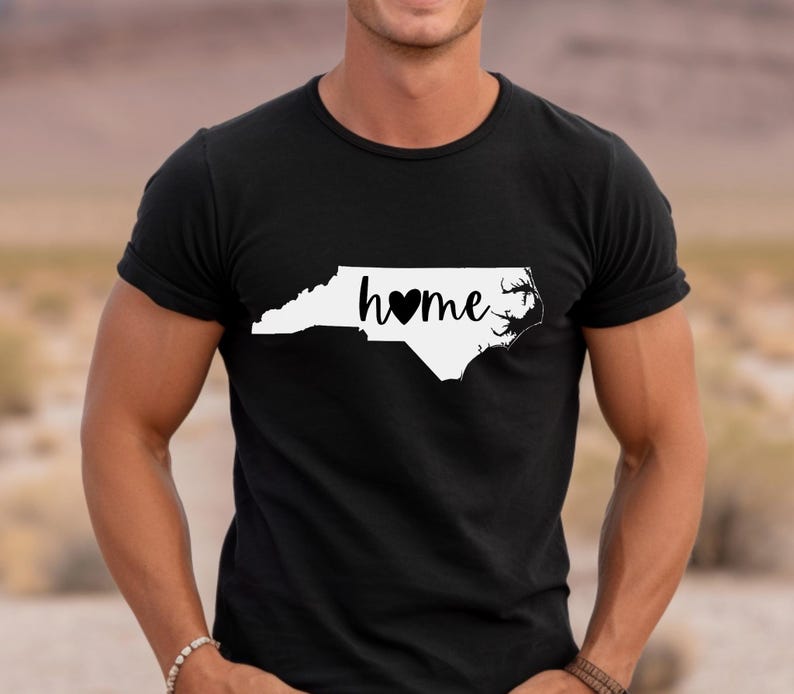 May include: Black t-shirt with a white outline of North Carolina and the word "home" with a black heart. The shirt is made of a soft material and has short sleeves.