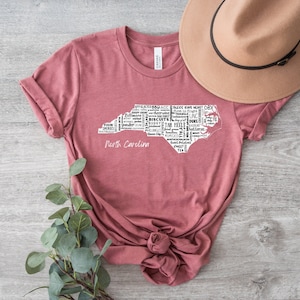 Pu&ograve; includere: Una maglietta color marsala con un contorno bianco dello stato della Carolina del Nord. Il contorno &egrave; riempito con parole relative alla Carolina del Nord, come "BBQ", "Tar Heels", "OBX" e "Biscuits". Le parole "North Carolina" sono stampate sotto il contorno.