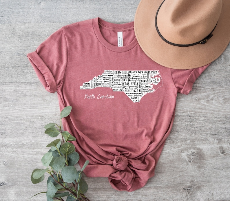 Pu&ograve; includere: Una maglietta color marsala con un contorno bianco dello stato della Carolina del Nord. Il contorno &egrave; riempito con parole relative alla Carolina del Nord, come "BBQ", "Tar Heels", "OBX" e "Biscuits". Le parole "North Carolina" sono stampate sotto il contorno.