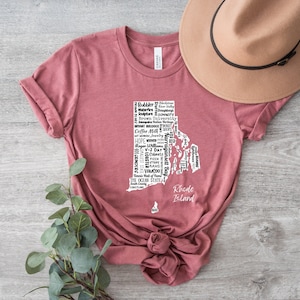 K&ouml;nnte beinhalten: Ein maronfarbenes T-Shirt mit einer wei&szlig;en Umrandung des Bundesstaates Rhode Island. Die Umrandung ist mit Text gef&uuml;llt, der sich auf Rhode Island bezieht, darunter "Coffee Milk", "Ocean State", "Hope", "Providence", "Newport" und "Block Island". Der Text "Rhode Island" steht unterhalb der Umrandung.