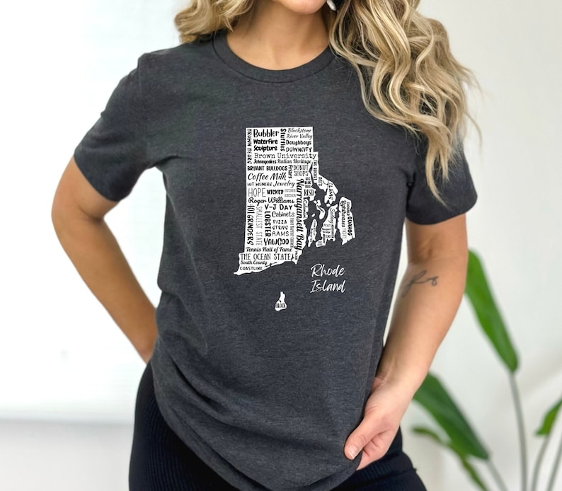 K&ouml;nnte beinhalten: Ein dunkelgraues T-Shirt mit einer wei&szlig;en Umrandung des Bundesstaates Rhode Island. Die Umrandung ist mit Text gef&uuml;llt, der sich auf Rhode Island bezieht, darunter "Coffee Milk", "Narragansett Bay", "Roger Williams" und "The Ocean State".