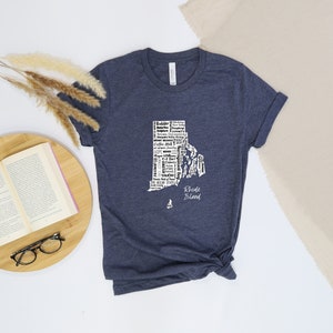 K&ouml;nnte beinhalten: Ein marineblaues T-Shirt mit einer wei&szlig;en Umrandung des Bundesstaates Rhode Island. Die Umrandung ist mit W&ouml;rtern gef&uuml;llt, die mit dem Bundesstaat in Verbindung stehen, darunter "Ocean State", "Newport", "Providence" und "Block Island". Die Worte "Rhode Island" stehen unter der Umrandung.