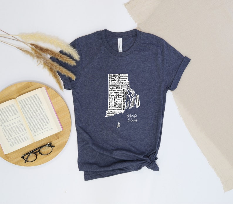 K&ouml;nnte beinhalten: Ein marineblaues T-Shirt mit einer wei&szlig;en Umrandung des Bundesstaates Rhode Island. Die Umrandung ist mit W&ouml;rtern gef&uuml;llt, die mit dem Bundesstaat in Verbindung stehen, darunter "Ocean State", "Newport", "Providence" und "Block Island". Die Worte "Rhode Island" stehen unter der Umrandung.
