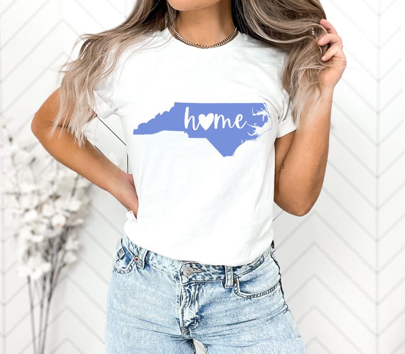 May include: White t-shirt with a light blue outline of North Carolina and the word "home" in a script font with a heart symbol. The shirt is made of a soft material, suitable for casual wear.