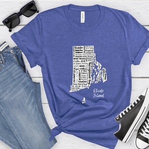 K&ouml;nnte beinhalten: Ein blaues T-Shirt mit einer wei&szlig;en Umrandung des Bundesstaates Rhode Island. Die Umrandung ist mit W&ouml;rtern gef&uuml;llt, die mit dem Bundesstaat in Verbindung stehen, wie "Coffee Milk", "WaterFire" und "The Ocean State".