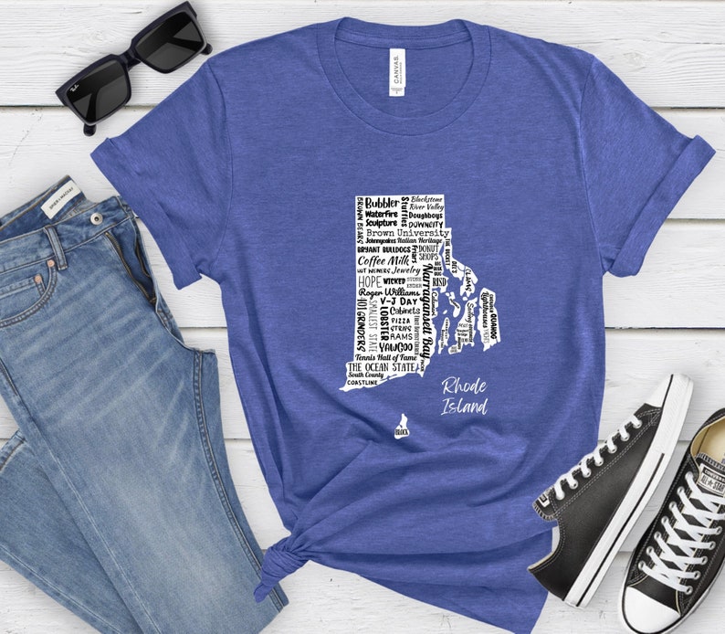 K&ouml;nnte beinhalten: Ein blaues T-Shirt mit einer wei&szlig;en Umrandung des Bundesstaates Rhode Island. Die Umrandung ist mit W&ouml;rtern gef&uuml;llt, die mit dem Bundesstaat in Verbindung stehen, wie "Coffee Milk", "WaterFire" und "The Ocean State".