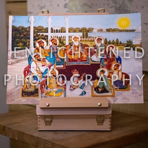 May include: A vibrant religious painting showing multiple figures in traditional dress, set against a lake and a bright sun. The artwork is displayed on a small wooden easel, with the words "ENLIGHTENED PHOTOGRAPHY" visible.