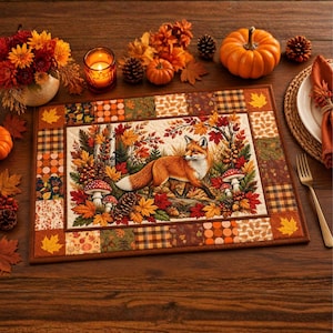 May include: A rectangular quilted placemat featuring a central illustration of a red fox surrounded by autumn leaves, pinecones, and mushrooms. The border consists of a patchwork pattern with squares of plaid, polka dots, and fall foliage motifs.