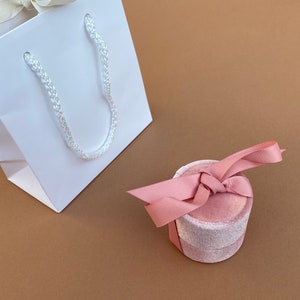 K&ouml;nnte beinhalten: Eine wei&szlig;e Geschenkt&uuml;te mit einem wei&szlig;en Seilhenkel und eine rosafarbene Samt-Ringbox mit einem rosafarbenen Satinband.