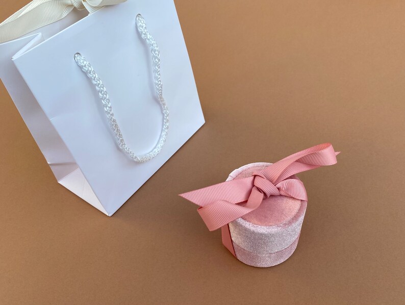 K&ouml;nnte beinhalten: Eine wei&szlig;e Geschenkt&uuml;te mit einem wei&szlig;en Seilhenkel und eine rosafarbene Samt-Ringbox mit einem rosafarbenen Satinband.
