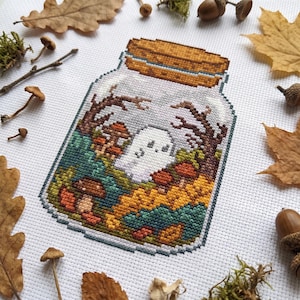 以下が含まれることがあります： 白い布に施されたクロスステッチ刺繍で、小さな白い幽霊、茶色のキノコ、裸の木の枝が入ったガラス瓶が描かれています。瓶には質感のある茶色の蓋があり、散らばった秋の葉を背景に配置されています。