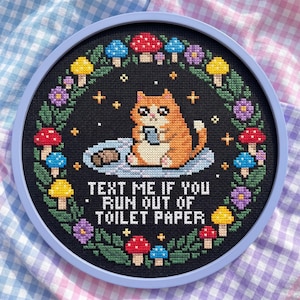 May include: Cross-stitch embroidery in a round frame. The design features an orange cat holding a phone, surrounded by mushrooms and flowers. Text reads: "TEXT ME IF YOU RUN OUT OF TOILET PAPER."