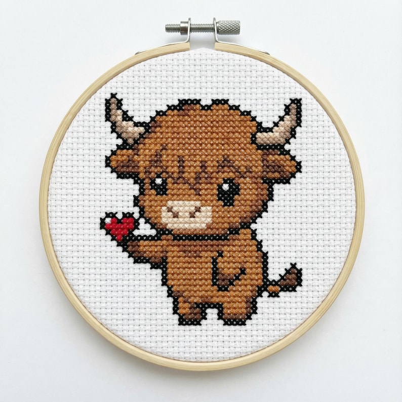 Puede incluir: Bordado de punto de cruz de una vaca de las Tierras Altas de dibujos animados sosteniendo un coraz&oacute;n rojo. La vaca es marr&oacute;n con ojos y cuernos negros, sobre un fondo blanco. El bordado est&aacute; enmarcado en un aro de madera redondo.
