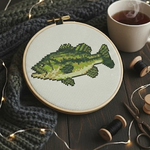 May include: A cross-stitch embroidery hoop featuring a green and yellow fish design. The embroidery is set against a light background, framed by a wooden hoop. The scene includes a cup of tea, thread spools, and scissors.