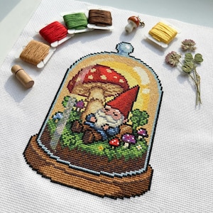 May include: Cross-stitch art featuring a gnome in a terrarium with a red-capped mushroom. The scene is surrounded by colorful flowers and greenery. Spools of thread in various colors and a mushroom charm are arranged around the artwork.