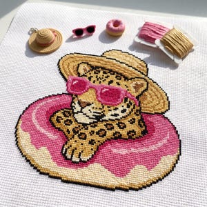 Puede incluir: Un bordado de punto de cruz de un leopardo con sombrero de paja y gafas de sol rosas, flotando en un flotador con forma de dónut rosa. Los accesorios incluyen un sombrero de paja en miniatura, gafas de sol rosas, un dónut y dos hilos.