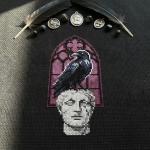 Puede incluir: Un diseño de punto de cruz que presenta un cuervo negro posado sobre un busto blanco, ambientado en una ventana gótica. El diseño está sobre un fondo negro, con plumas y amuletos decorativos. La paleta de colores incluye negro, blanco y tonos morados.