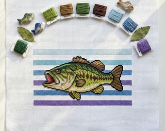May include: Cross-stitch of a largemouth bass with an open mouth, set against blue and purple horizontal stripes. The image is surrounded by spools of thread in various colors, small fish figurines, and green leaves. The background is white.