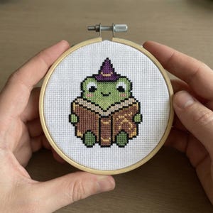 Könnte beinhalten: Kreuzstich-Stickerei einer grünen Froschfigur mit lila Hexenhut, die ein braunes Buch liest. Der Frosch befindet sich auf weißem Stoff, der von einem hölzernen Stickrahmen umgeben ist. Ein handgefertigtes Kunsthandwerk.