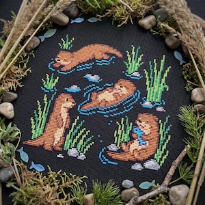 以下が含まれることがあります： 黒い布に施されたクロスステッチ刺繍で、泳いだり青い魚を持ったりしている4匹の茶色のカワウソが描かれています。周囲には緑の水生植物、青い水の波紋、小さな灰色の石が配置されています。