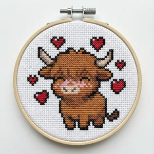 May include: A cross-stitch embroidery in a wooden hoop features a brown Highland cow with black outlines and a pink muzzle. Red hearts surround the cow on a white background. The embroidery is secured with a metal screw.