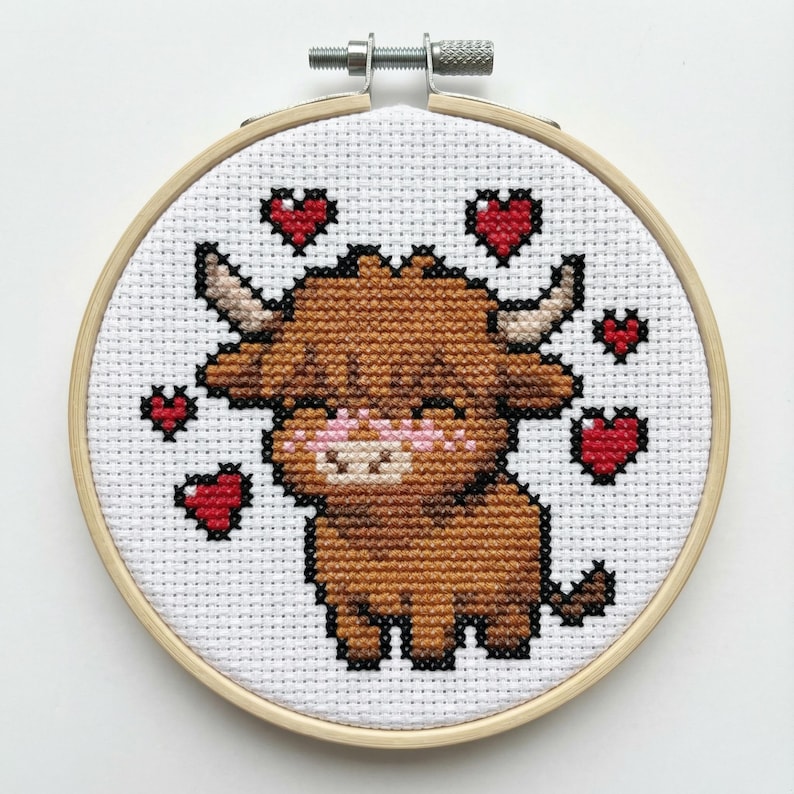 May include: A cross-stitch embroidery in a wooden hoop features a brown Highland cow with black outlines and a pink muzzle. Red hearts surround the cow on a white background. The embroidery is secured with a metal screw.