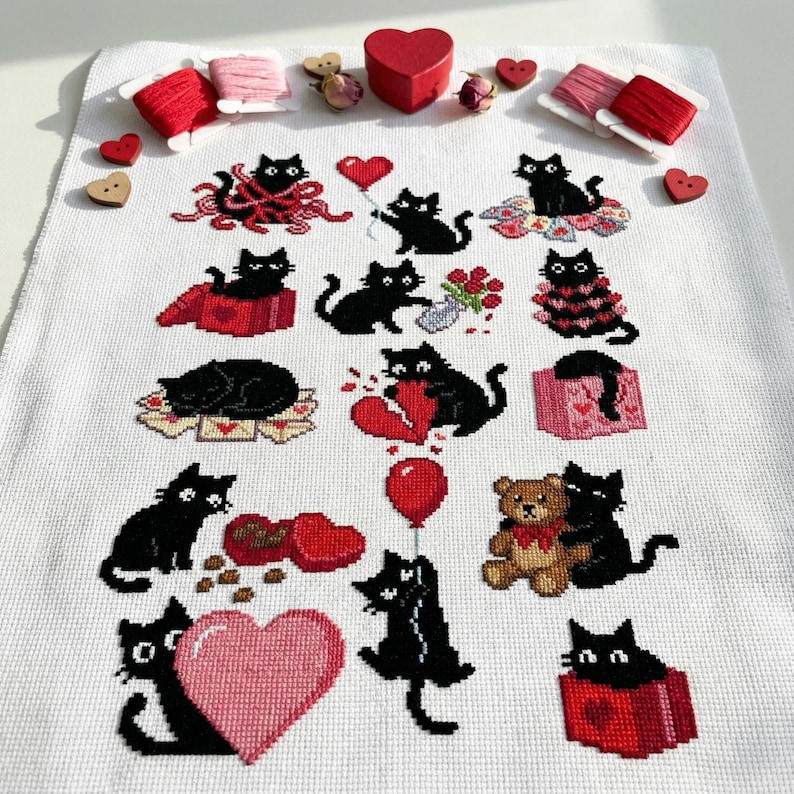 May include: A white cross-stitch fabric featuring various black cat designs with Valentine's Day themes. The cats are depicted with hearts, gifts, and balloons. Red and pink thread spools and wooden heart accents are also present.