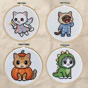 May include: Four cross-stitch hoops featuring adorable cat designs. One cat is dressed as a fairy with a wand, another in pajamas, a third in a pumpkin costume, and the last in a dinosaur outfit. Each design is set in a wooden hoop.