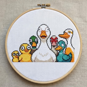 Puede incluir: Un bordado de punto de cruz con un grupo de patos de dibujos animados en un aro de madera. Los patos son de varios colores, incluyendo amarillo, verde, blanco, naranja y turquesa. El tejido es blanco.