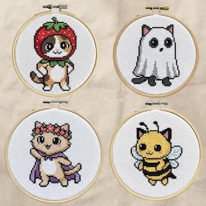 May include: Four cross-stitch embroidery hoops featuring cat designs. One cat is dressed as a strawberry, another as a ghost, a third with a flower crown and cape, and the last as a bee. Each design is set in a wooden hoop.