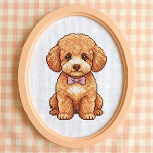 May include: A cross-stitch of a seated, light brown dog with a pink bow tie, framed in a peach-colored oval. The dog has large, floppy ears and a white patch on its chest. The background is a white fabric with a peach gingham pattern.