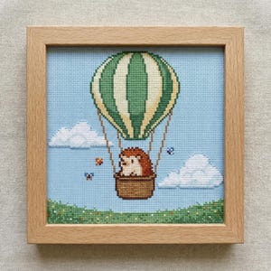 Puede incluir: Arte de punto de cruz de un erizo en un globo aerostático con un diseño de rayas verdes y crema. La obra de arte está enmarcada en un marco de madera marrón claro, sobre un cielo azul claro con nubes blancas y mariposas.