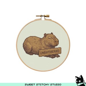 May include: A cross-stitch embroidery hoop featuring a brown capybara with closed eyes, holding a sign that reads "UNBOTHERED". The embroidery is set against a light green background, framed by a wooden hoop.