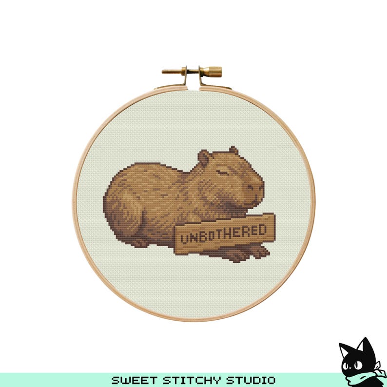 May include: A cross-stitch embroidery hoop featuring a brown capybara with closed eyes, holding a sign that reads "UNBOTHERED". The embroidery is set against a light green background, framed by a wooden hoop.