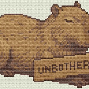 May include: A pixelated illustration of a capybara in shades of brown, lying down with its eyes closed. The capybara holds a sign that reads "UNBOTHERED" in block letters. The background is a light beige.